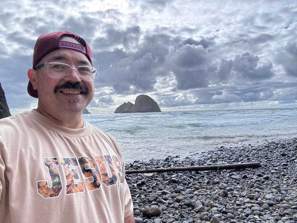 man wearing tan "JESUS" shirt on a rocky beach
