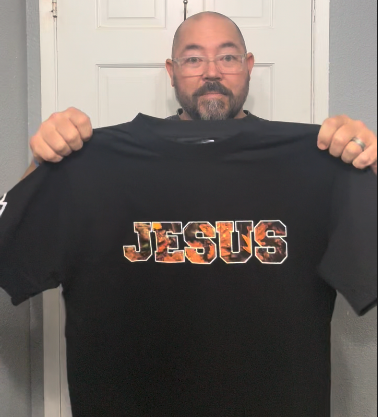 Man holding a black t-shirt with 'JESUS' printed on it in front of a white door.