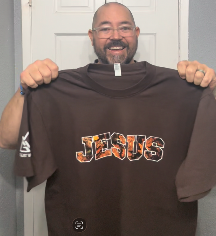 Man holding a brown t-shirt with 'JESUS' printed on it in front of a white door.