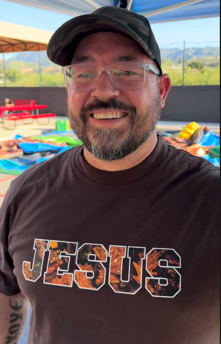 Man wearing a brown 'JESUS' t-shirt with a pool and outdoor area in the background.