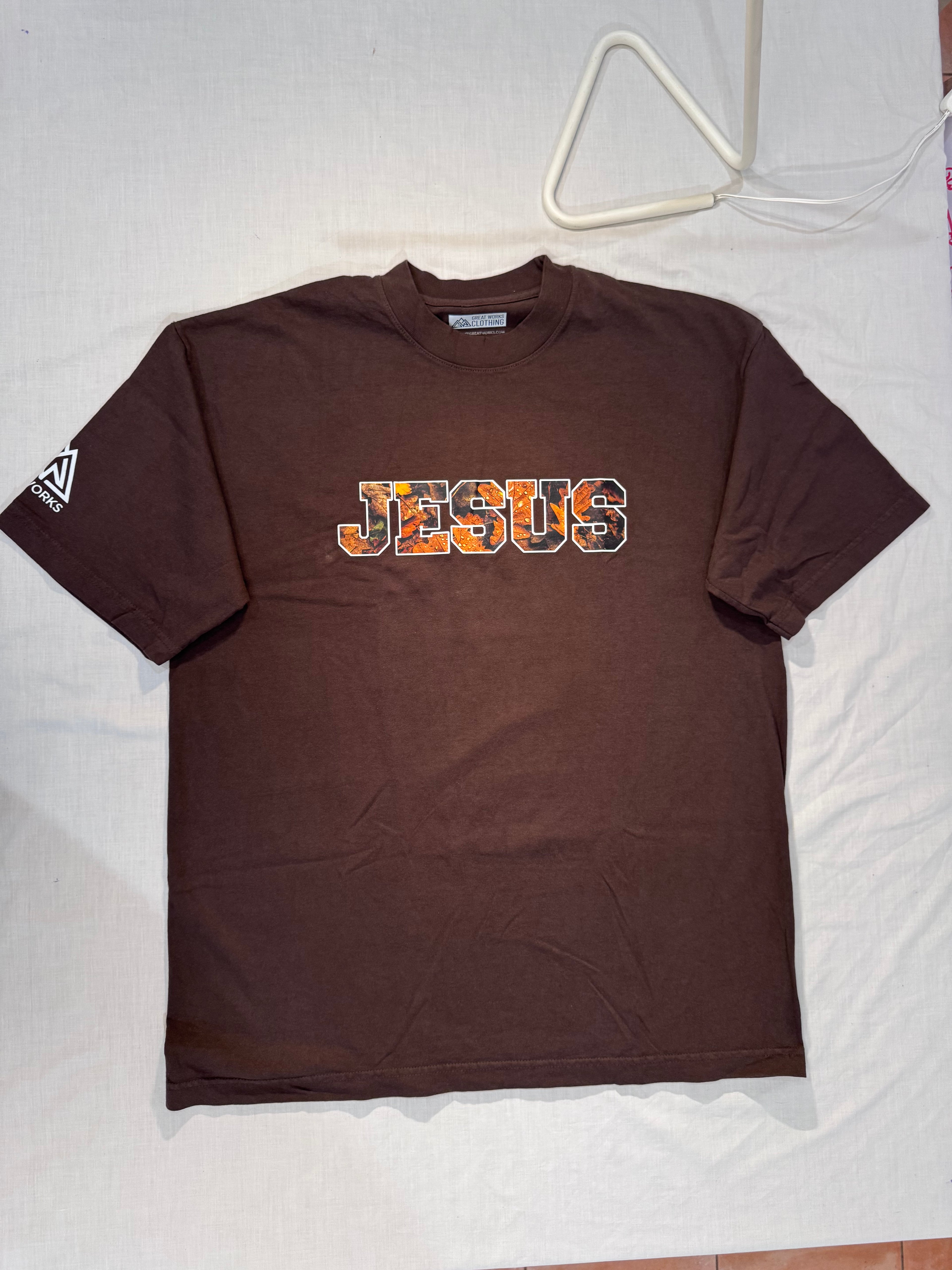 Brown t-shirt with 'JESUS' printed on it, displayed on a white surface.