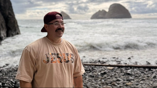 Person wearing a 'Jesus' t-shirt on a rocky beach with ocean and cliffs in the background