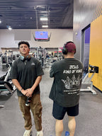 Two individuals in a gym setting, one wearing headphones and a black t-shirt with text, the other in a black t-shirt with a graphic design.