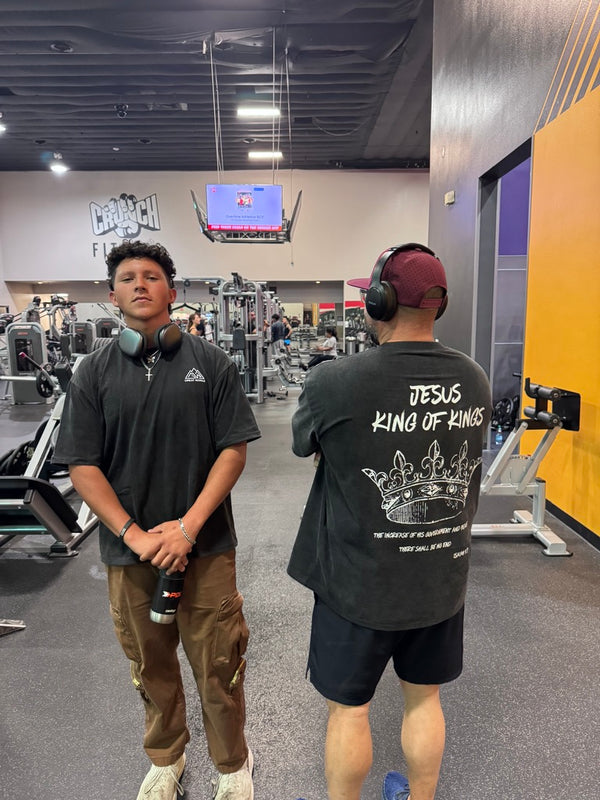 Two individuals in a gym setting, one wearing headphones and a black t-shirt with text, the other in a black t-shirt with a graphic design.